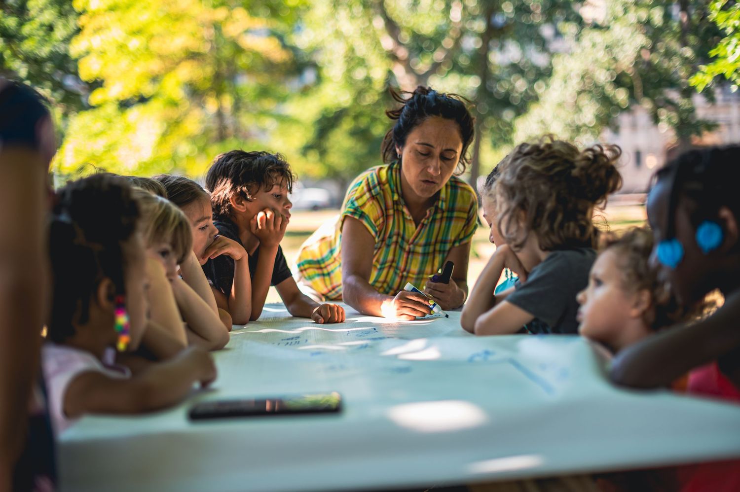 A group gathers around an outdoor table for an educational or recreational activity in natural lighting.