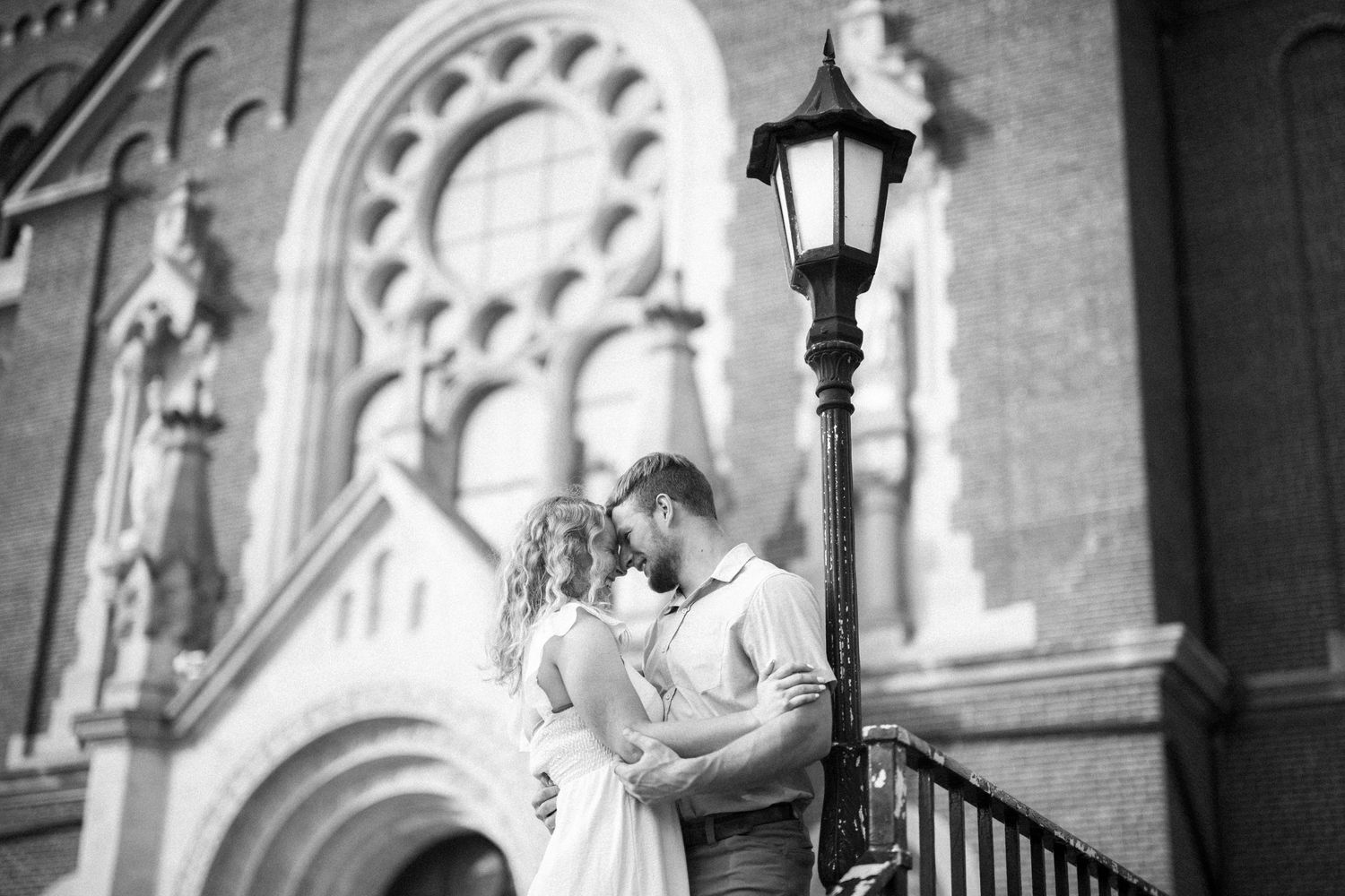 A romantic couple embrace on church steps next to an ornate lamp post in an elegant black and white photograph.