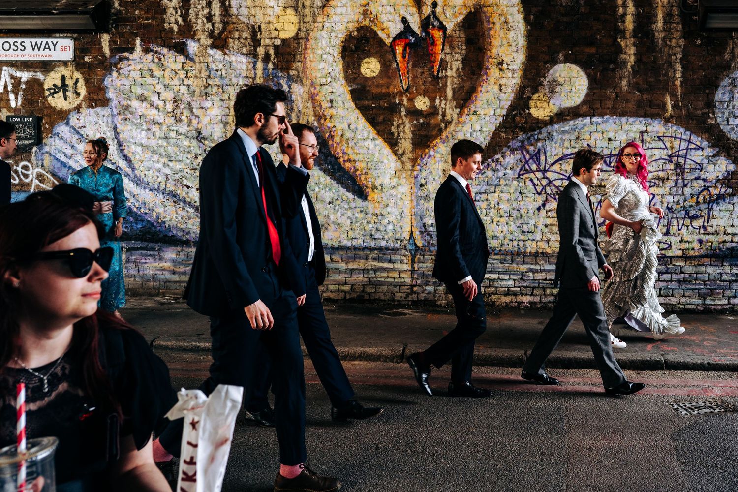 Wedding party walks past a colorful mosaic mural in an urban setting.