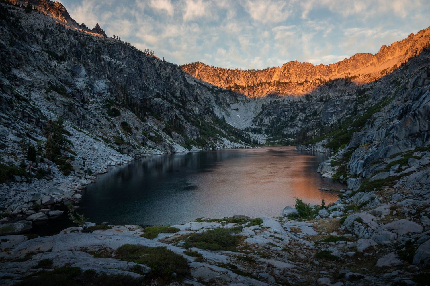 Alpine lake reflects golden sunset light on rocky mountain peaks in a remote wilderness setting.