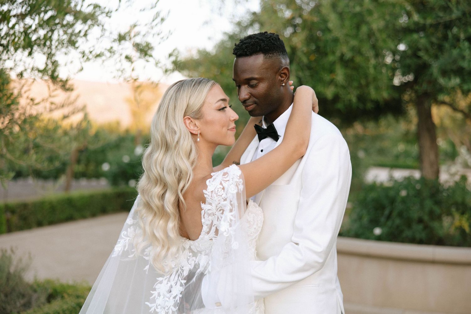 Bride in lace gown and groom in white tuxedo sharing romantic moment in garden at sunset.