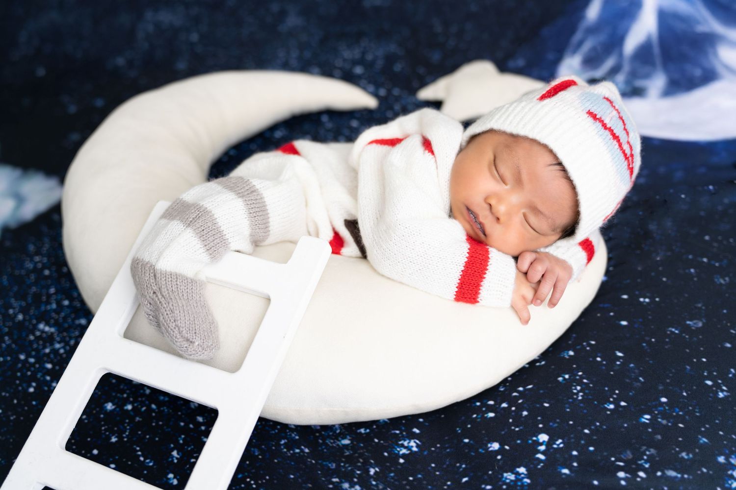 Sleeping baby wearing white and red outfit on a moon prop with stars background.