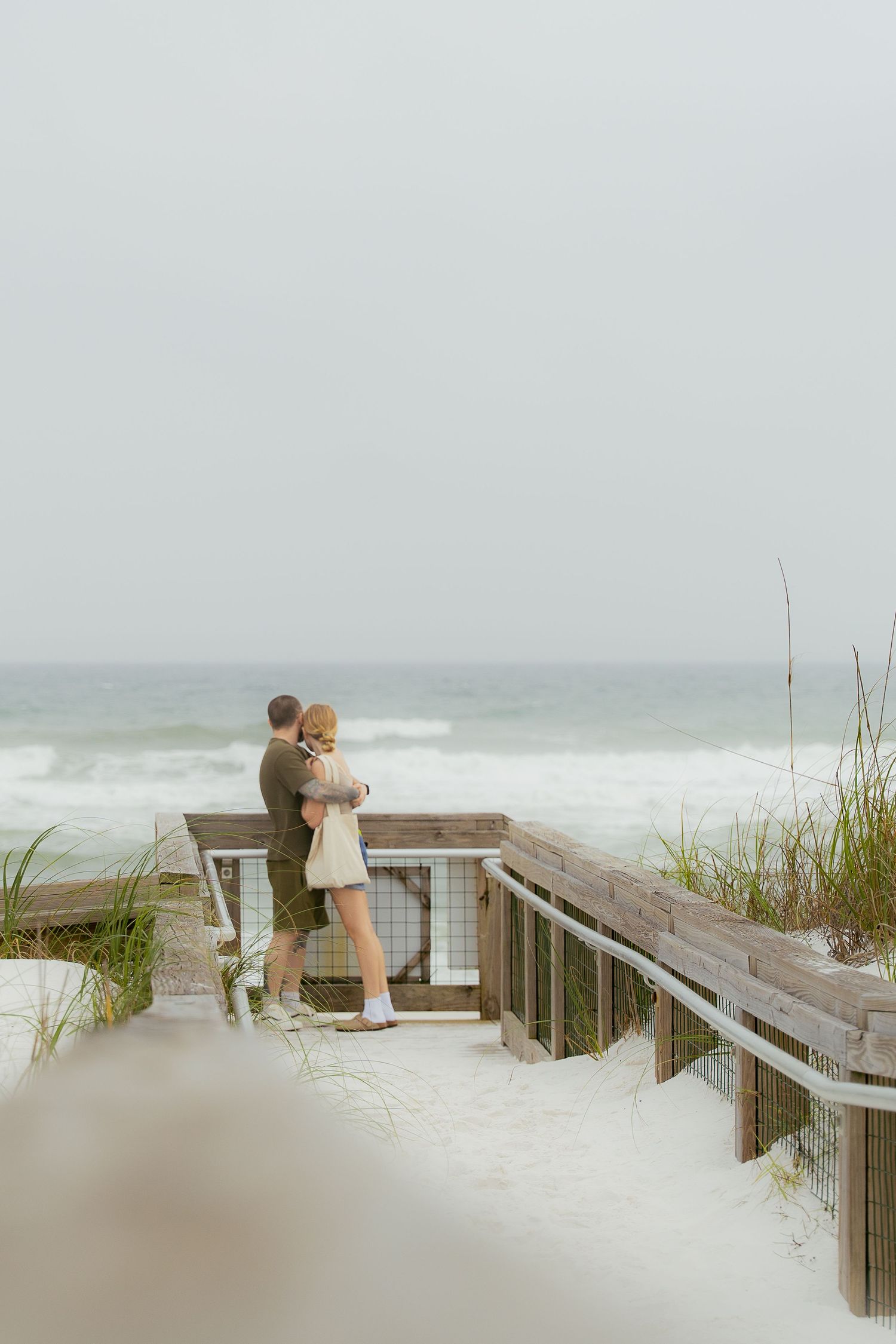 A couple embraces on a wooden boardwalk overlooking a misty beach with sea oats and waves in the background.