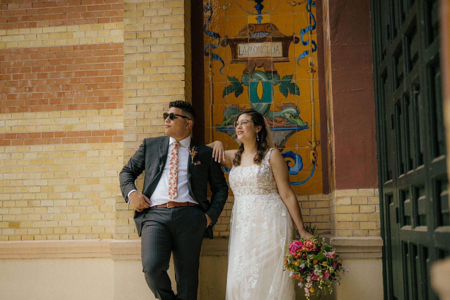 Two newlyweds pose casually against a brick wall with ornate doorway details, the bride holding a vibrant bouquet.