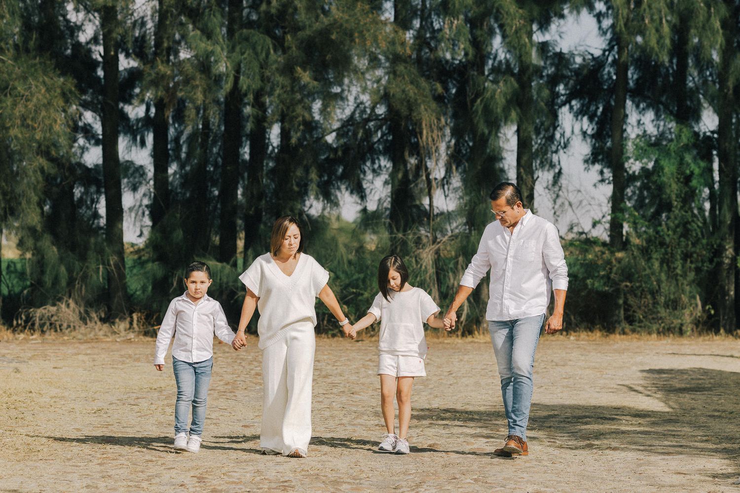 A family of four holding hands while walking together on a dirt path surrounded by tall evergreen trees.