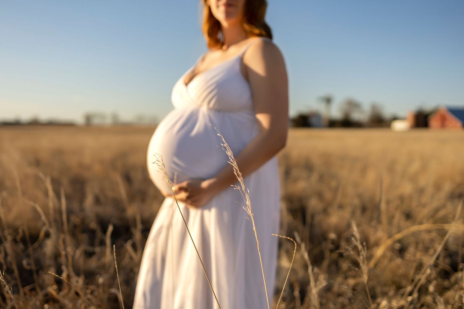 Silhouetted profile of an expectant mother in a white dress standing in a sunlit golden field at sunset.
