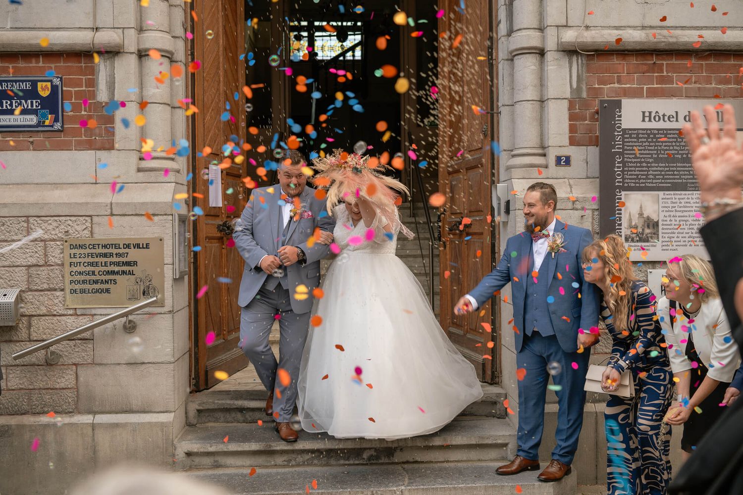 A couple exits a stone building while guests shower them with colorful confetti in a celebratory moment.