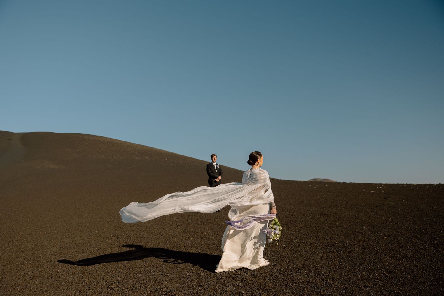 Person in flowing white dress stands on dark volcanic sand dune against blue sky as fabric billows dramatically in wind.