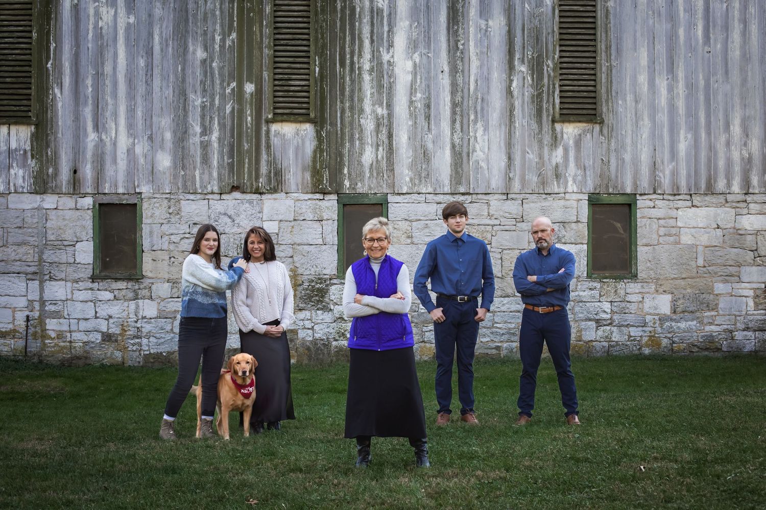 A group of people and a golden retriever pose together in front of a weathered stone building with green shutters.