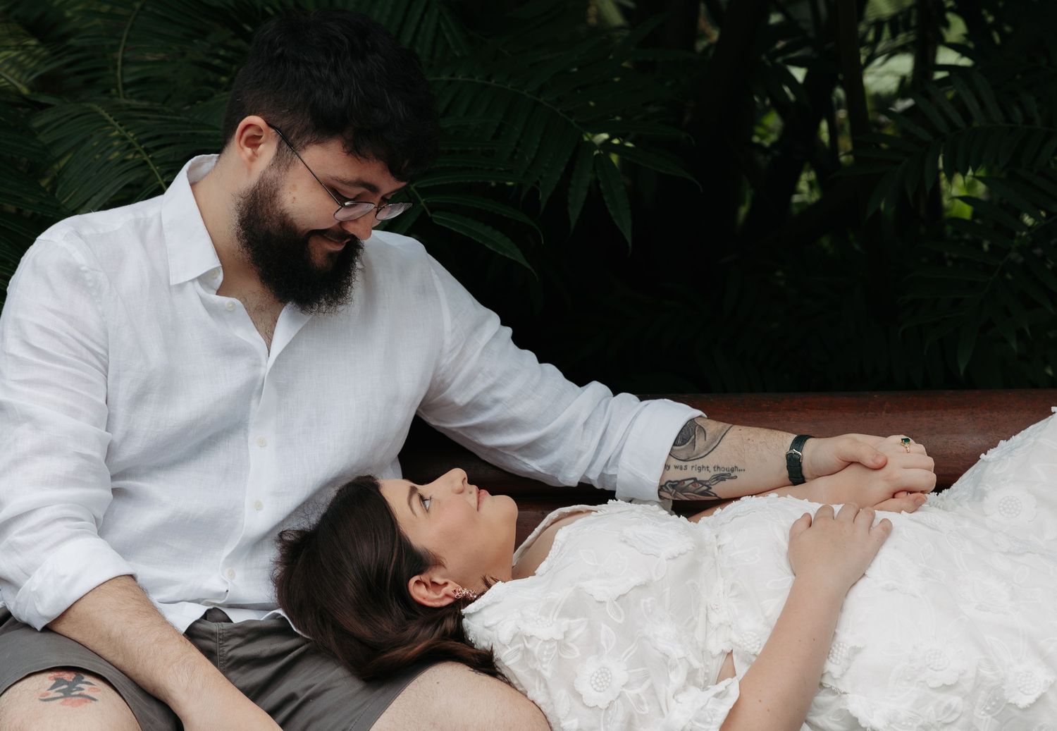 A couple in light colored clothing share a romantic moment on a path surrounded by lush green trees and bamboo.