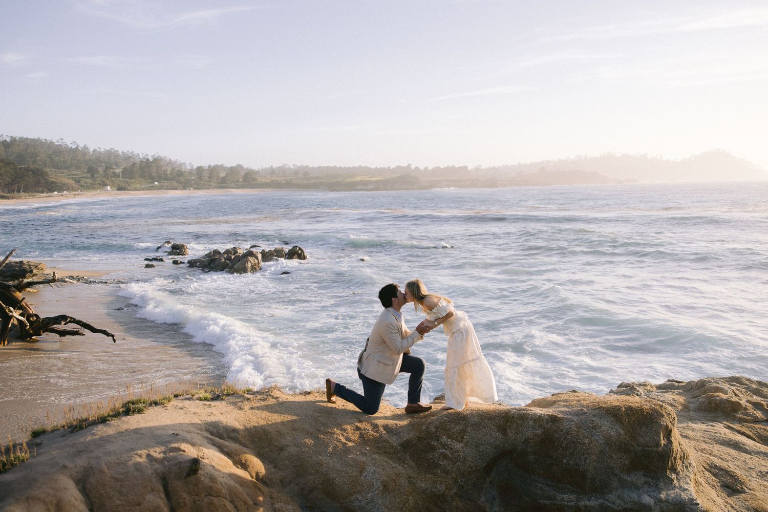 A couple shares an intimate moment on rocky coastal cliffs with crashing waves and a beach in the background.