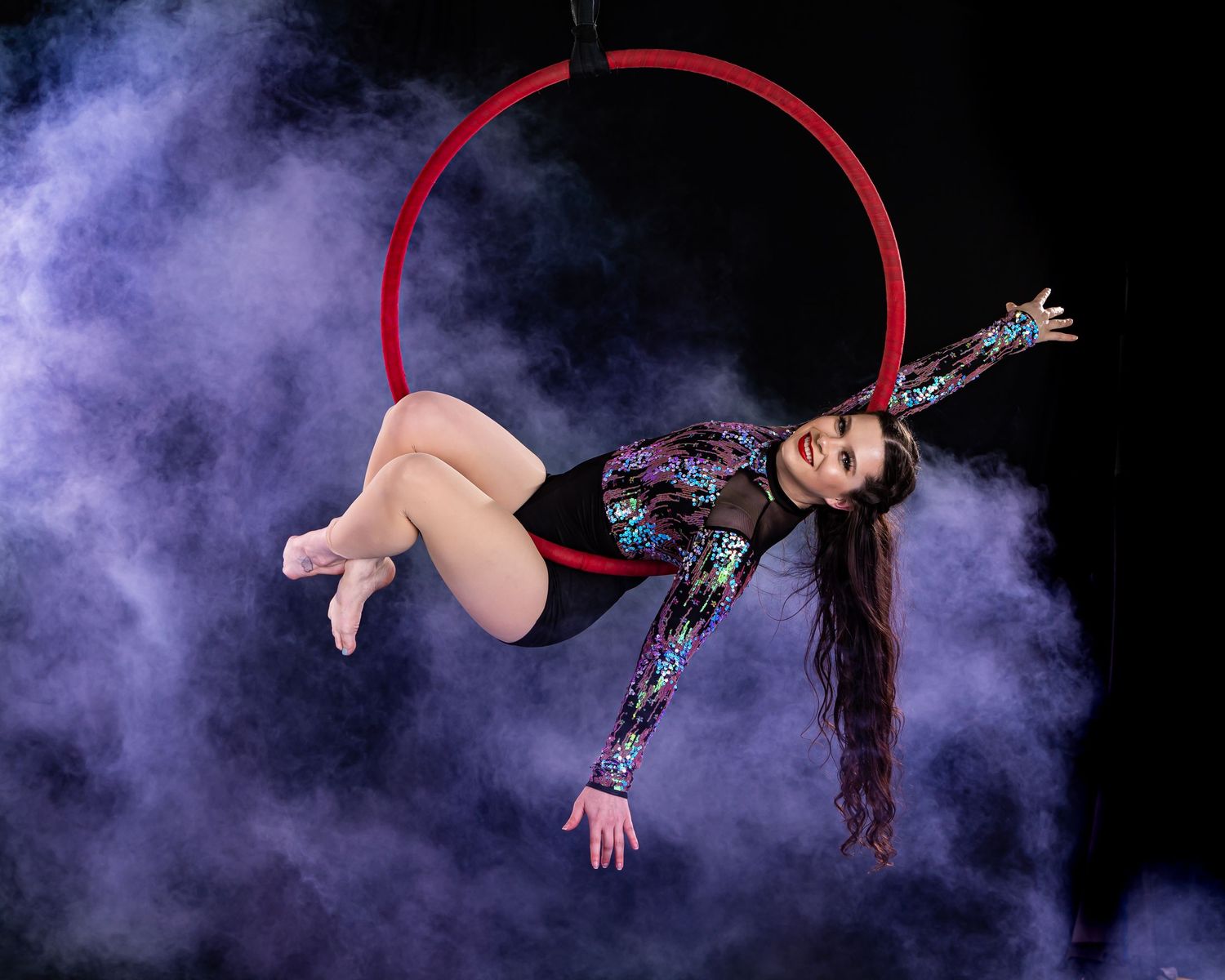 Aerial artist performs on red hoop against purple smoke backdrop.