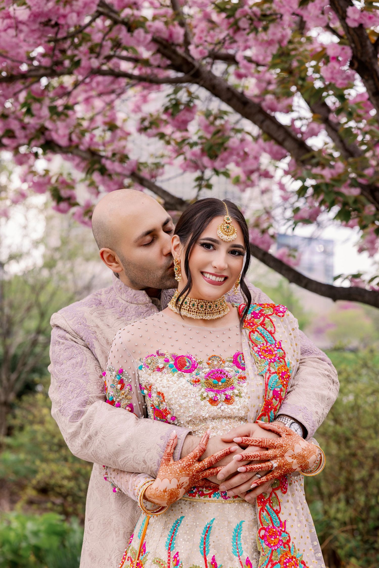 Traditional South Asian wedding couple embraces under blooming pink cherry blossom trees in spring garden setting.