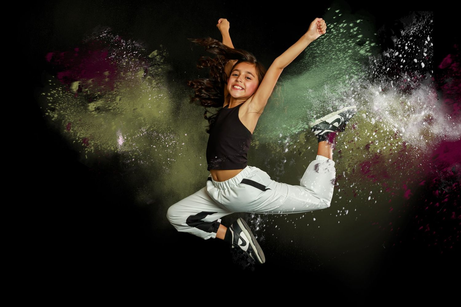 Dancer in black top and white pants leaps joyfully against colorful powder explosion background.