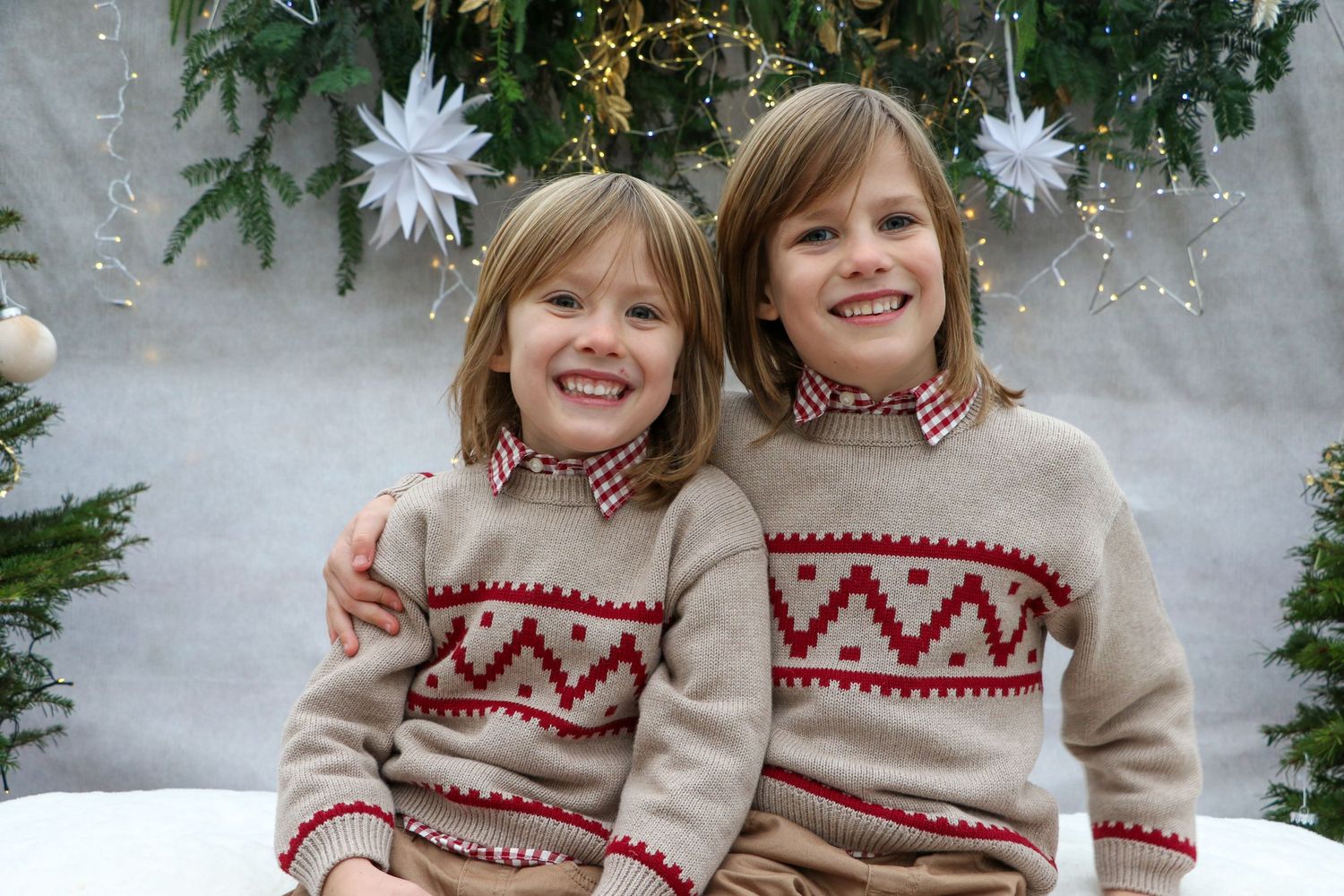 Two children wearing matching cream and red Nordic pattern sweaters in front of Christmas decorations.