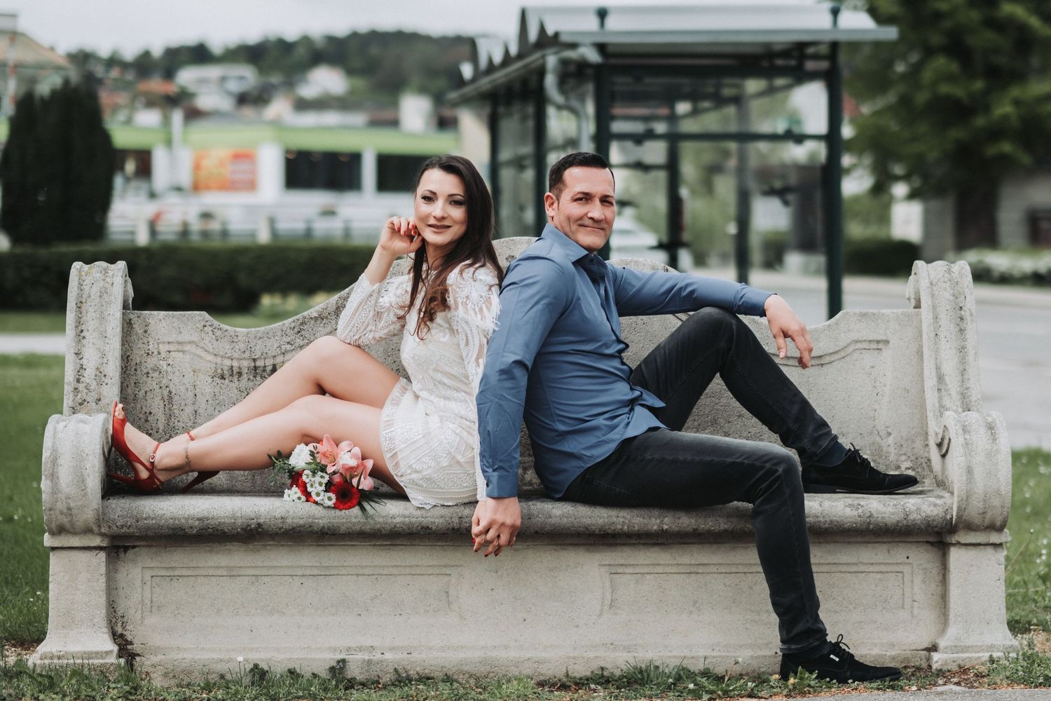 A couple sits casually on a stone bench in a garden setting with the woman in white and man in blue.