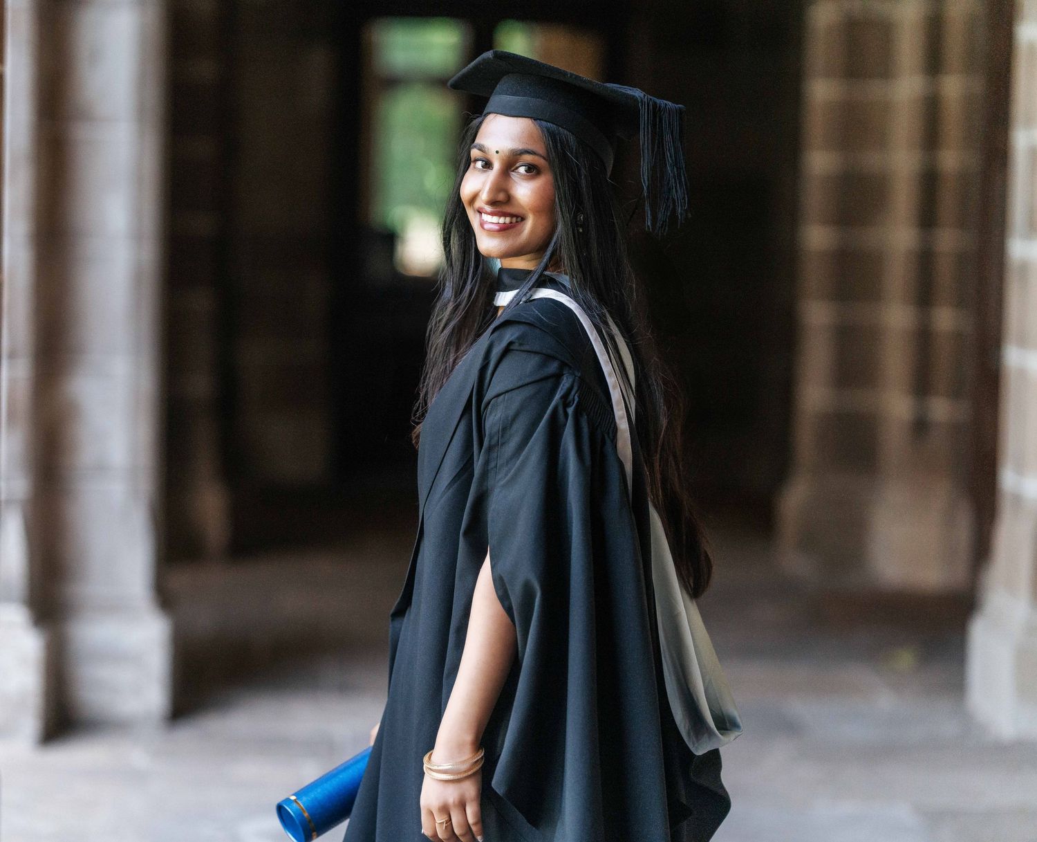 Graduate in traditional cap and gown poses in historic stone archway.