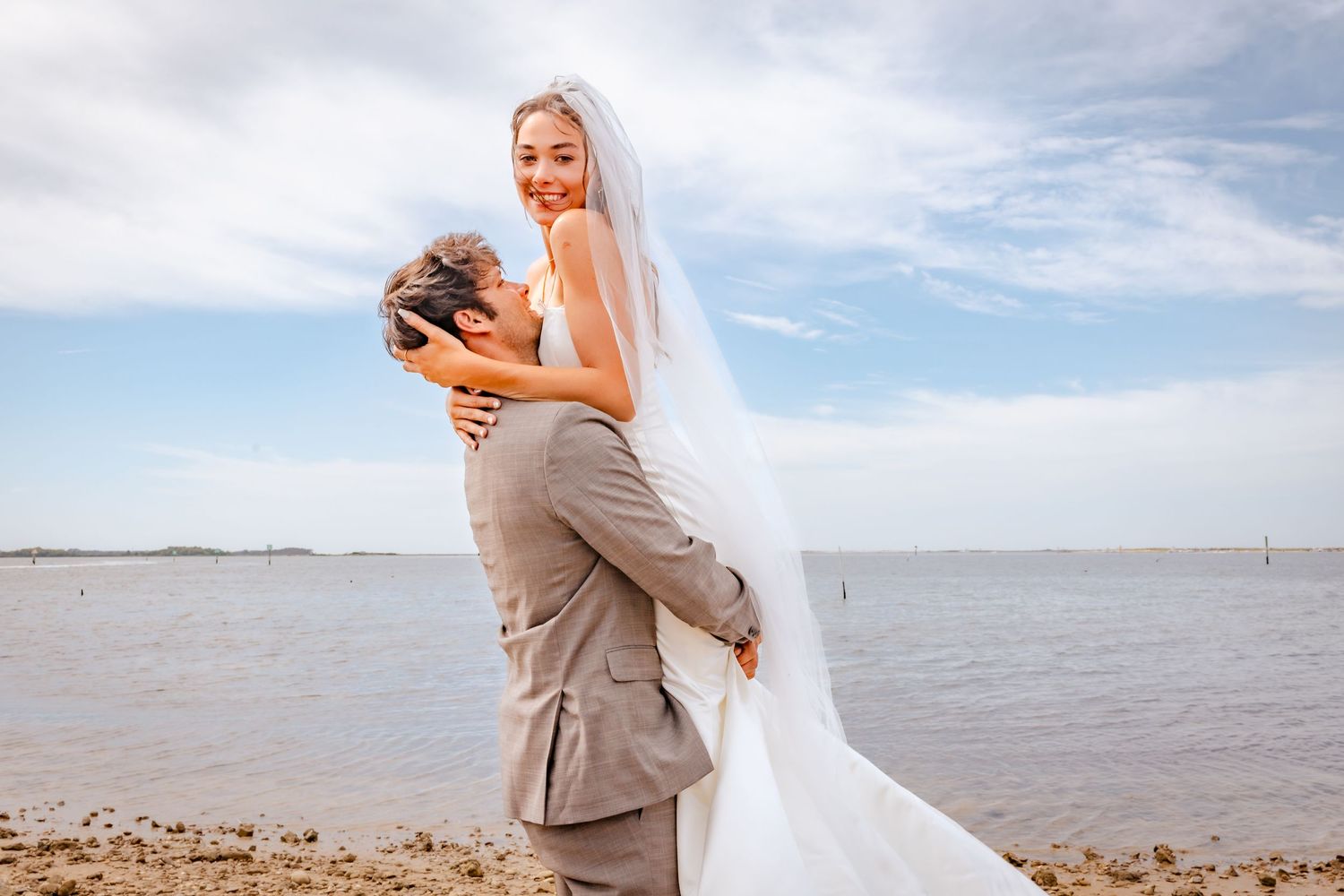 A couple celebrates their wedding day with a romantic embrace by the ocean under a bright blue sky.