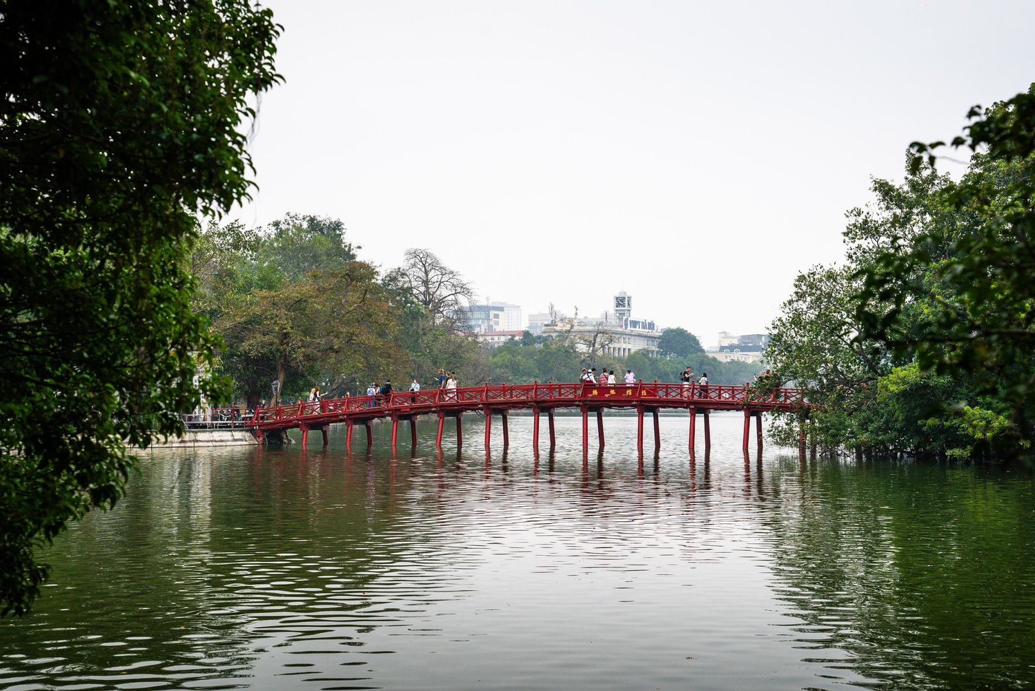 The iconic red Huc Bridge spans across Hoan Kiem Lake in Hanoi, Vietnam, surrounded by lush green trees.