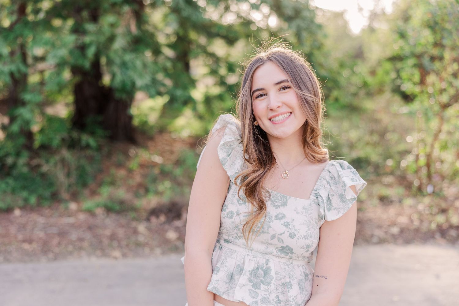 A person in a floral ruffle sleeve dress poses outdoors on a sunny day with greenery in the background.