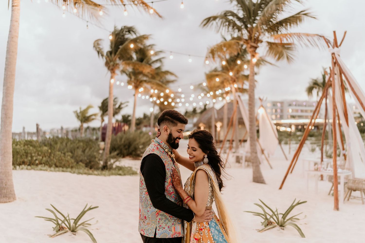 A couple shares a romantic moment near teepees and string lights under palm trees on a tropical beach.