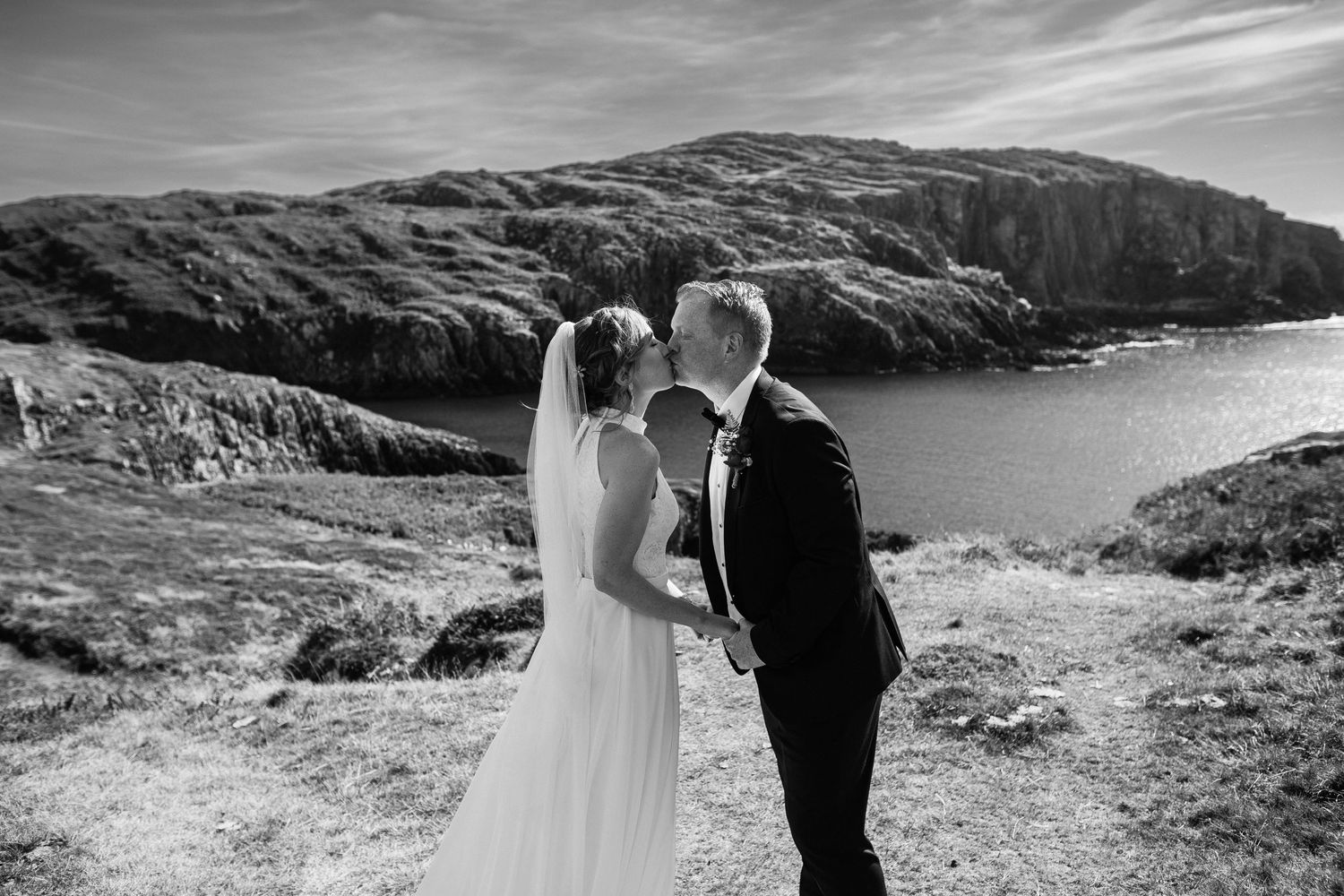 A wedding couple shares an intimate moment on a dramatic coastal cliff overlooking the ocean in black and white.