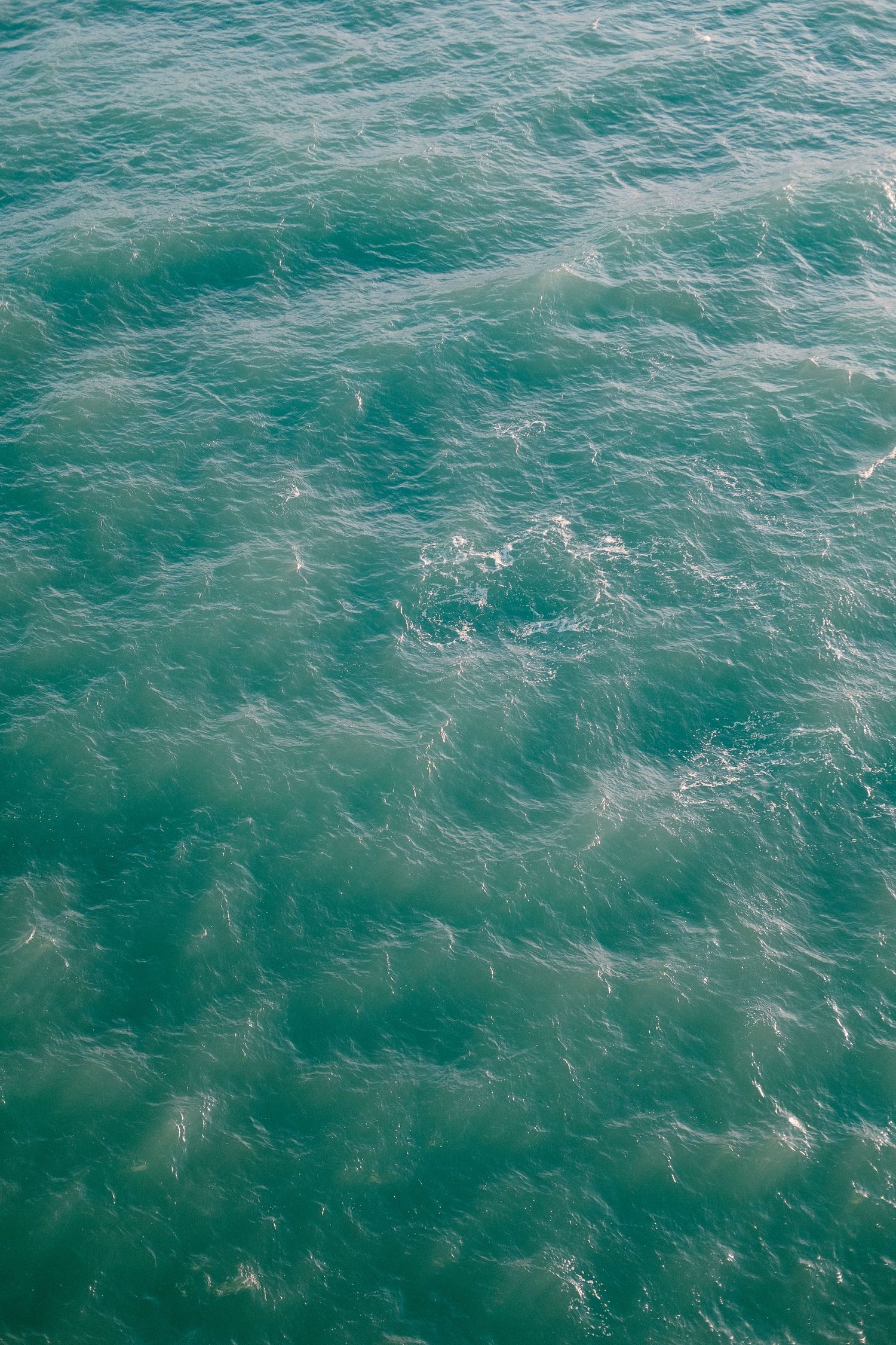 Aerial view of rippling turquoise ocean water with gentle waves and white foam patterns.