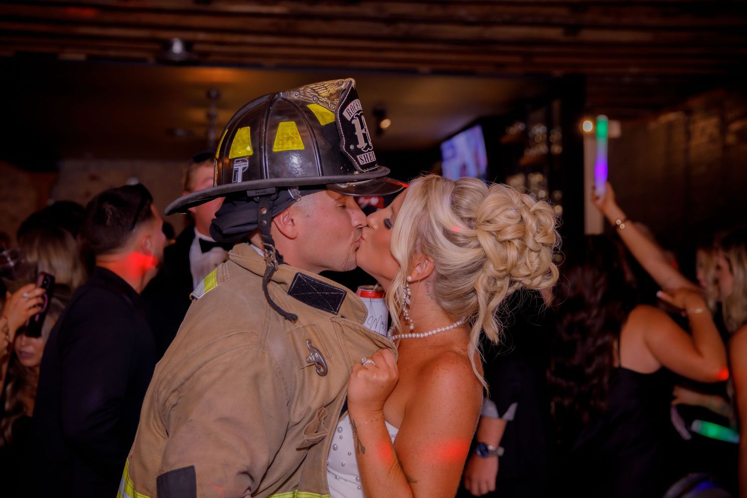 A couple embraces on a dance floor while one wears a firefighter uniform at a wedding reception.