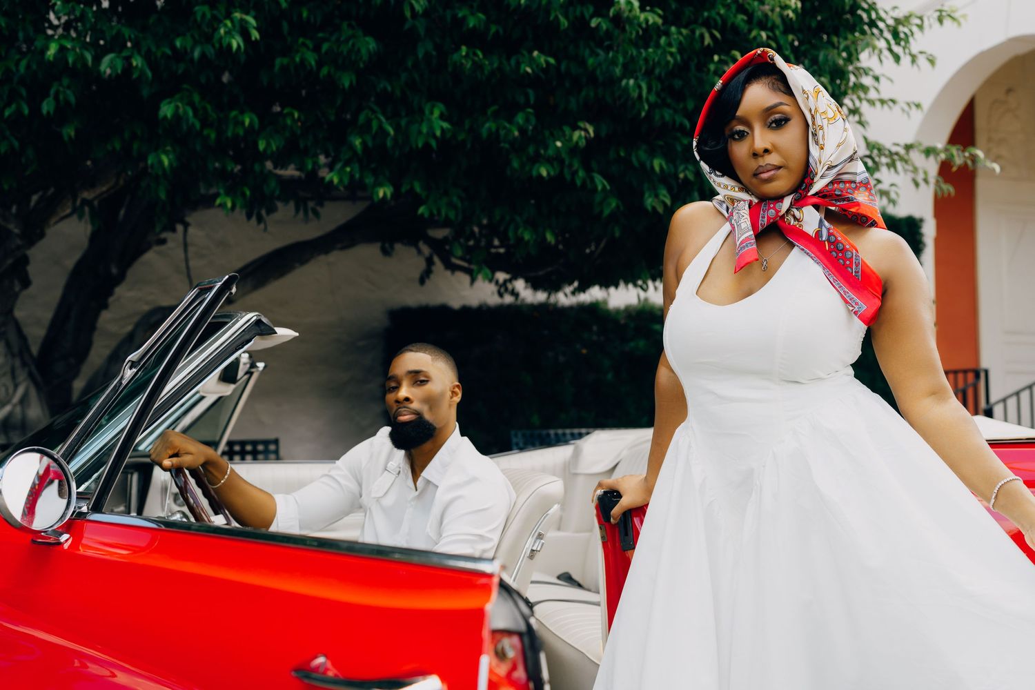 Vintage-style wedding photo with couple posing near red classic car, bride wearing white dress and red headscarf.