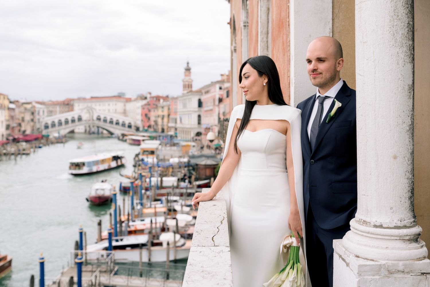 Couple in wedding attire poses on Venice balcony overlooking Grand Canal with Rialto Bridge in background.