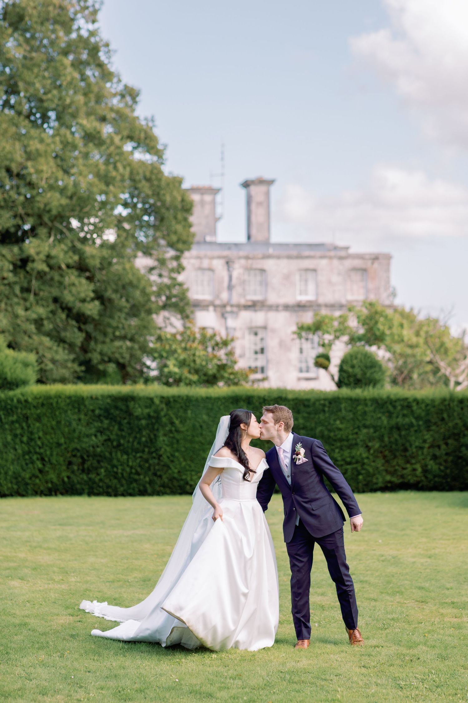 A romantic couple in wedding attire shares tender moments on a manicured lawn with a stately building in the background.