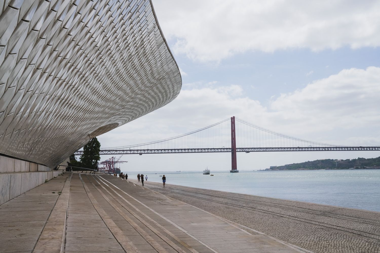 Modern MAAT museum facade in Lisbon with wooden promenade, visitors, and Ponte 25 de Abril bridge over Tagus River.