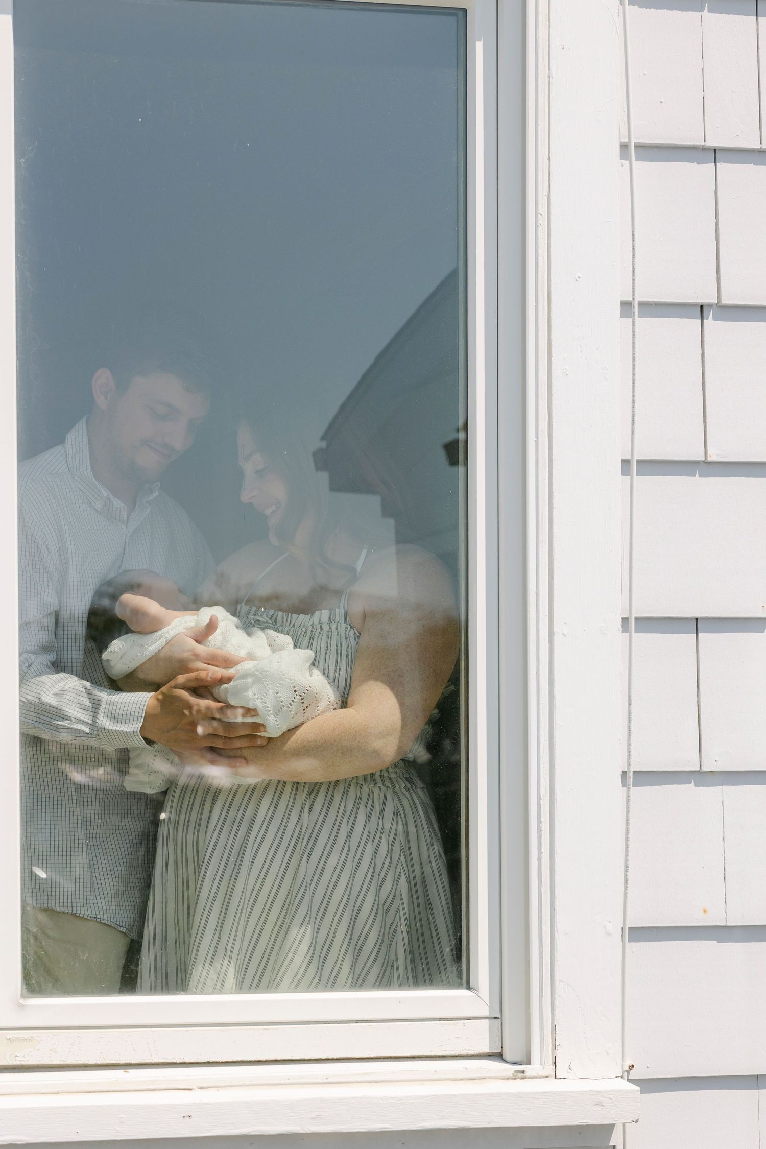 A couple stands by a window holding a newborn baby, viewed from outside through the glass pane.
