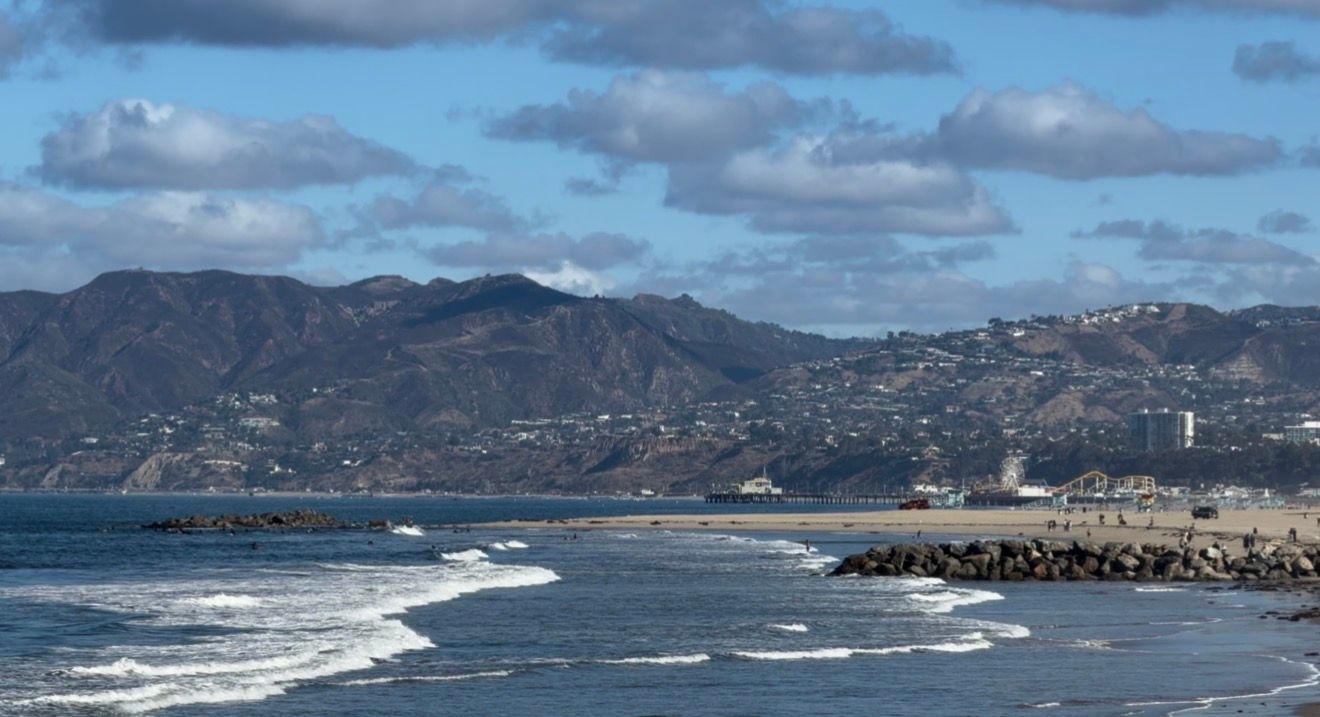 Scenic coastal view of Malibu beach with rocky jetty, ocean waves, and Santa Monica Mountains backdrop.