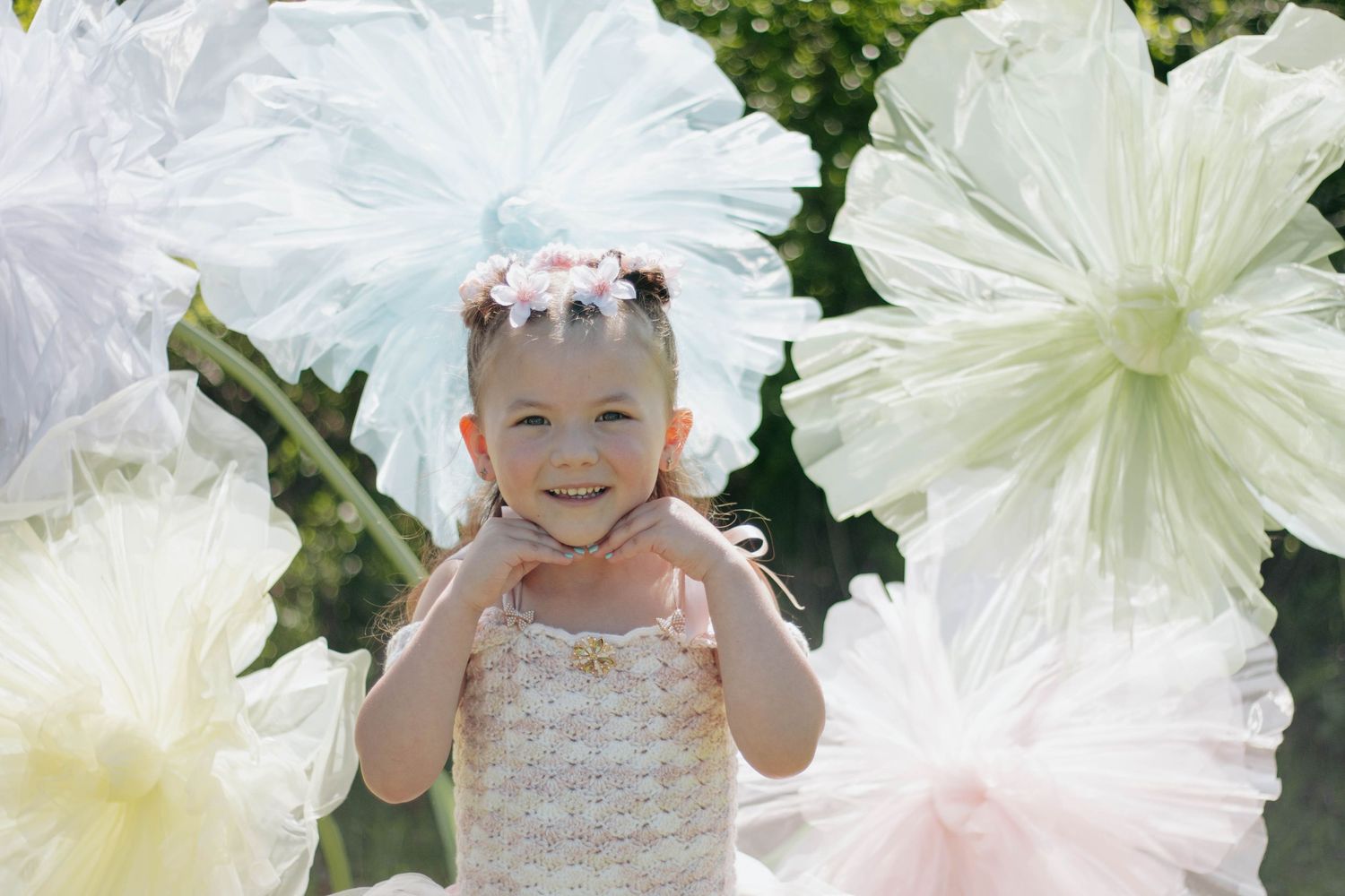 Young girl smiling in white dress surrounded by large decorative paper flowers outdoors.