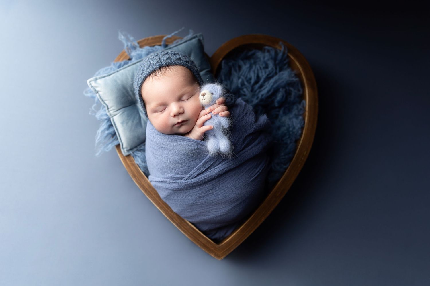 Newborn baby swaddled in blue wrap sleeping in wooden heart-shaped bowl with tiny stuffed animal and pillow.