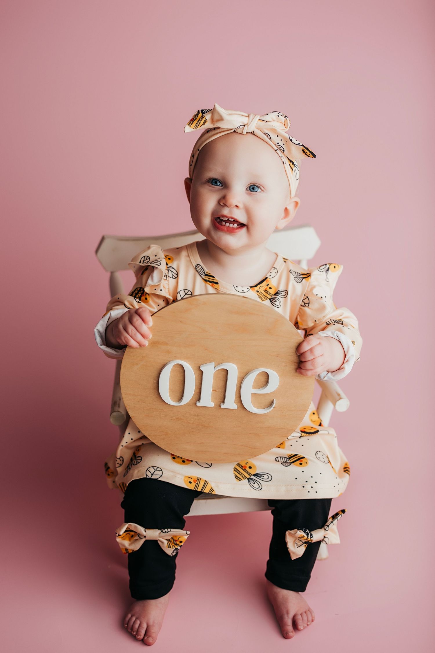 Smiling baby girl in bee-print outfit holds a wooden 'one' sign, celebrating her first birthday against a pink background.