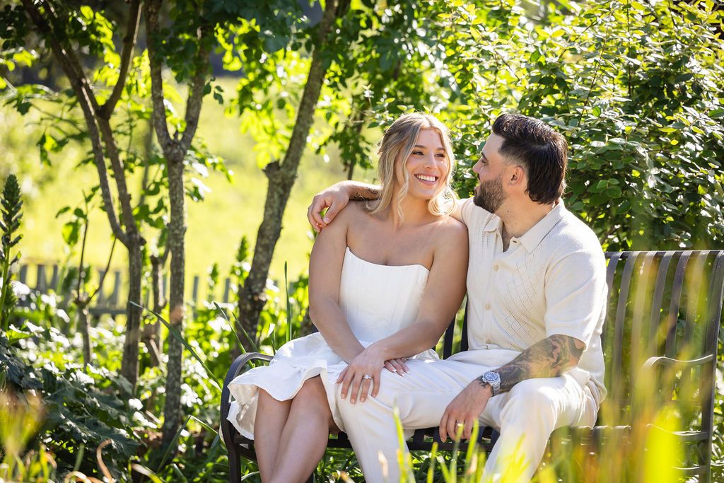 Couple sitting together on a bench surrounded by lush green trees, smiling and sharing a tender moment outdoors.