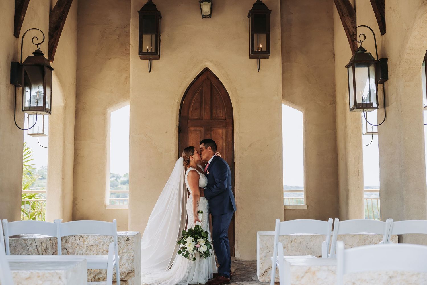 Bride and groom share a kiss at a rustic chapel entrance with arched wooden doors and elegant lanterns.