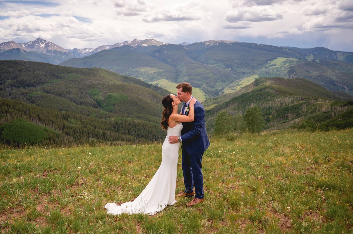 Bride and groom share a kiss on a scenic mountain meadow with stunning Rocky Mountain peaks in the background.
