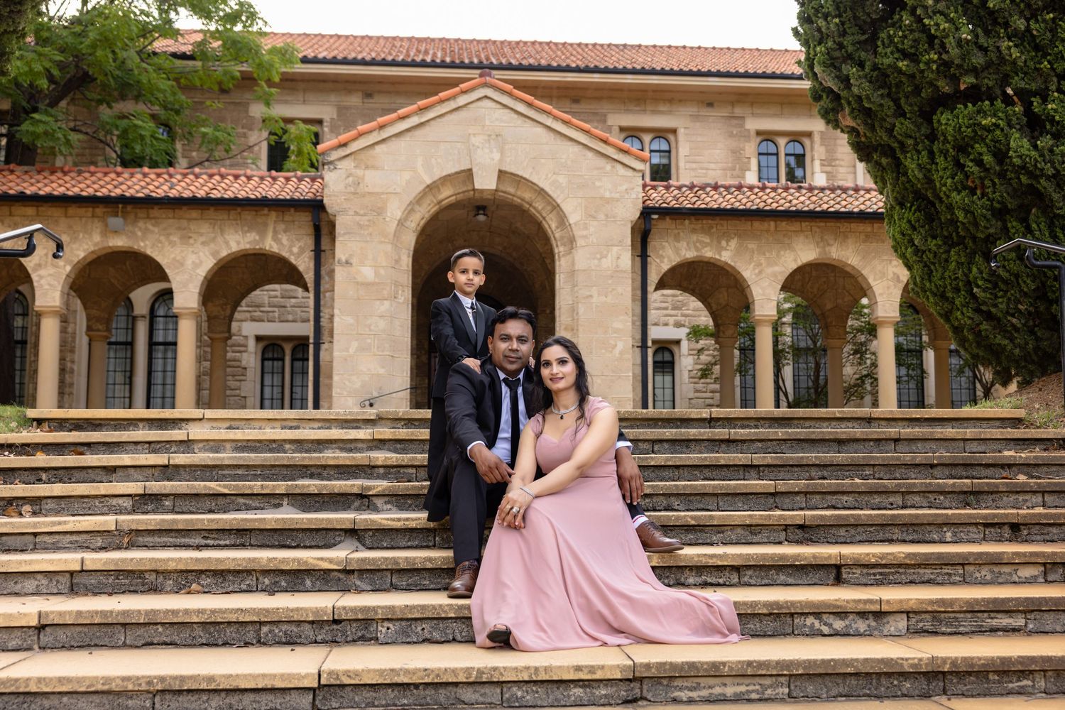 Family of three posing on stone steps before an arched building, woman in pink gown, man and child in suits.