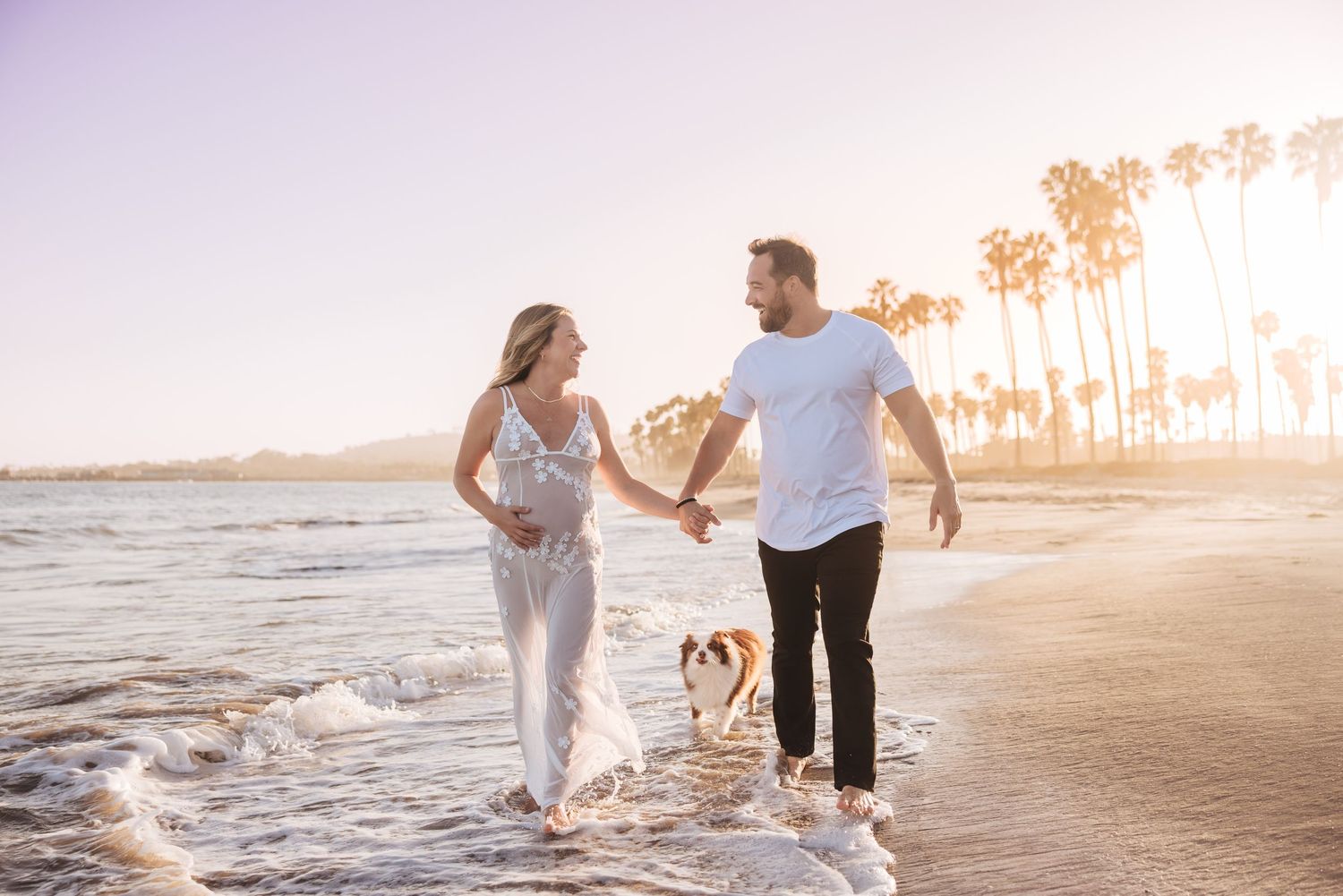 Couple holding hands walking on beach at sunset with dog and palm trees in background Santa Barbara maternity photographer