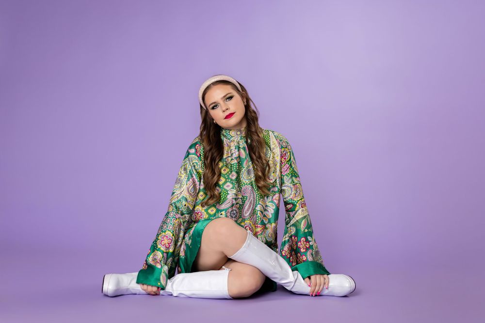 Model wearing a vibrant green floral dress with puff sleeves and embroidered details poses against purple backdrop.