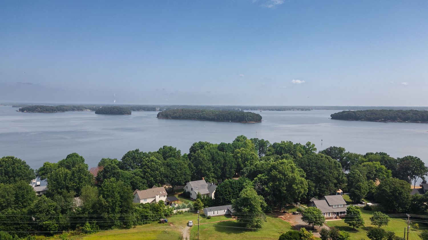 Aerial view of a lakeside community surrounded by lush green trees with scenic islands and calm blue water in the background.