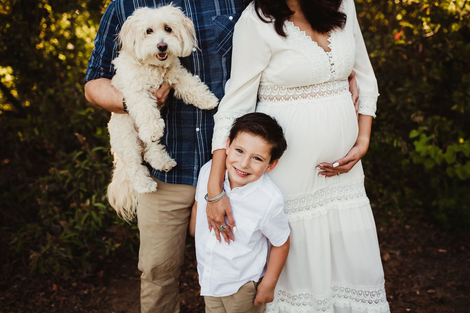 Pregnant woman in white dress with smiling boy, man holding fluffy white dog during outdoor maternity photo session.