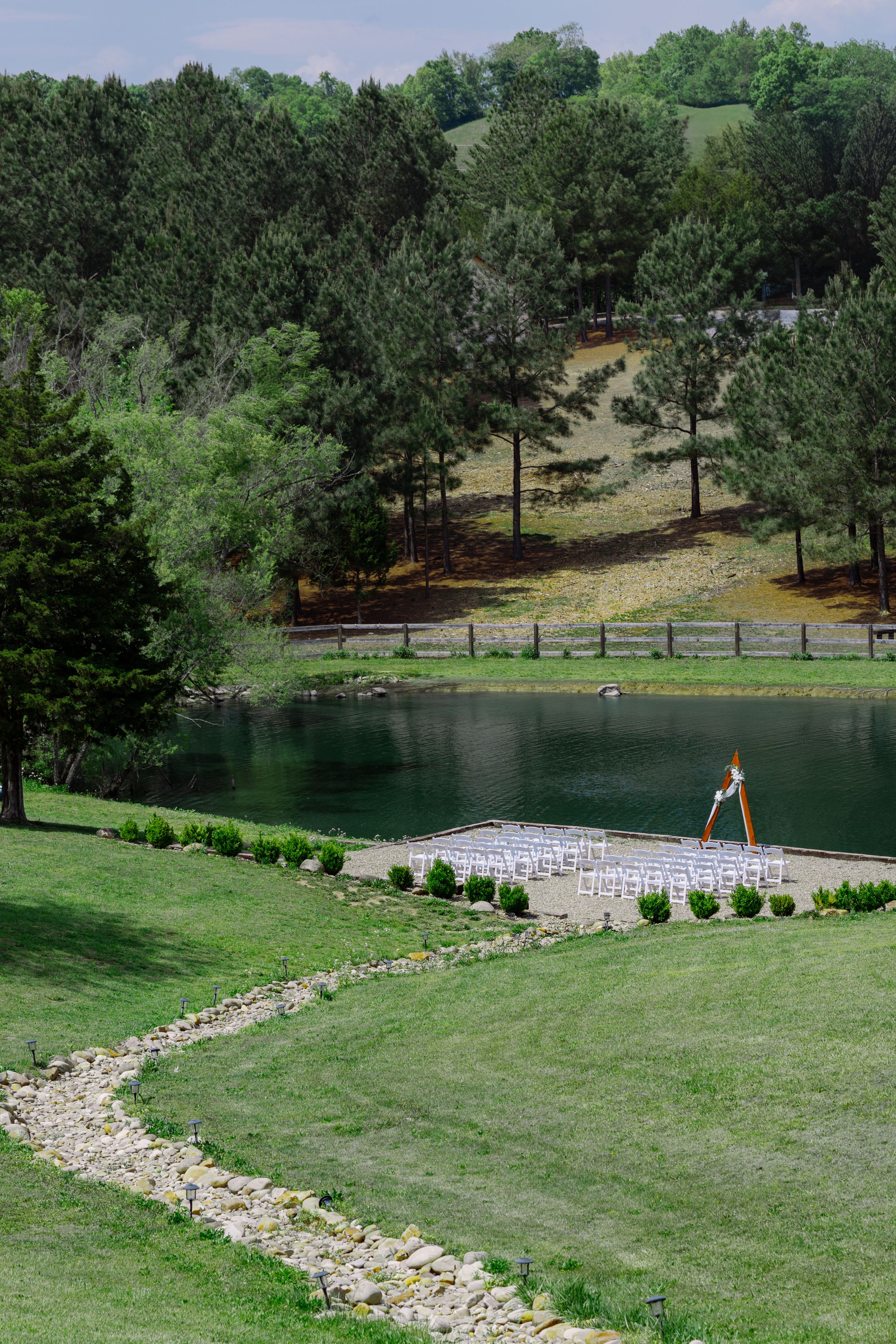 Outdoor wedding ceremony setup with white chairs beside a serene pond, surrounded by lush green trees and rolling hills.