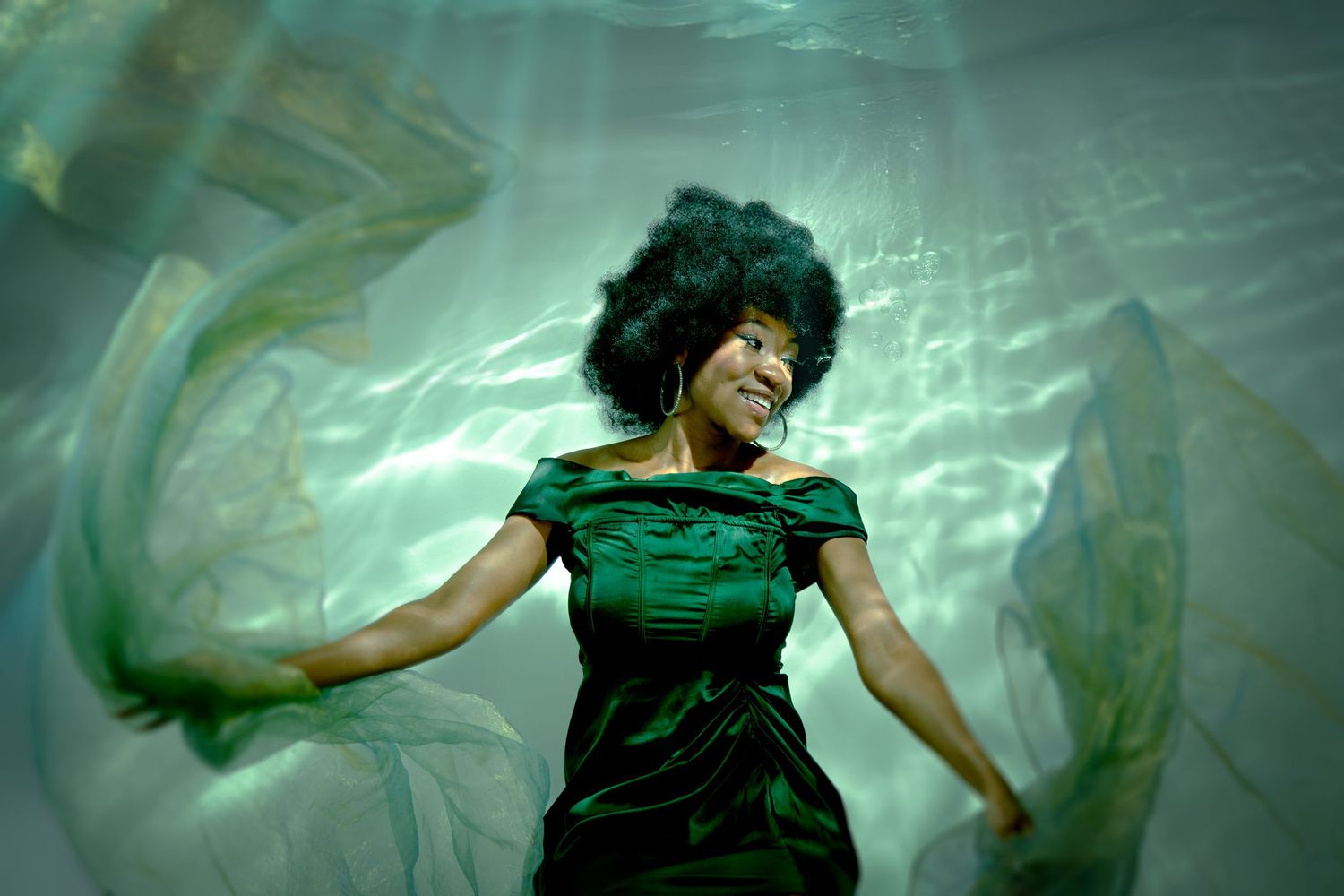 Woman in green dress underwater, surrounded by flowing fabric with dappled light creating ethereal effect.