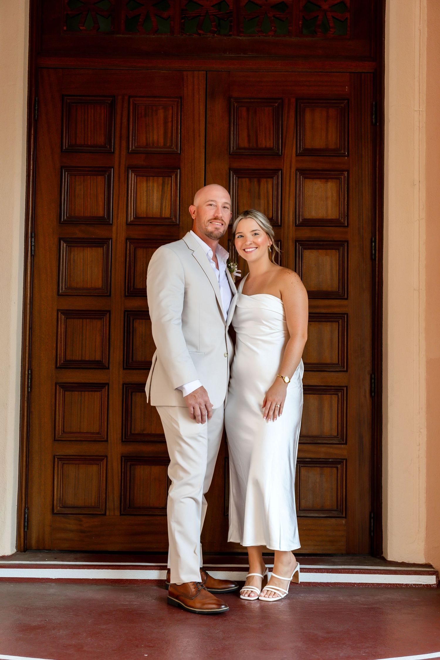 A couple in wedding attire poses before large wooden doors, man in tan suit, woman in white satin midi dress.