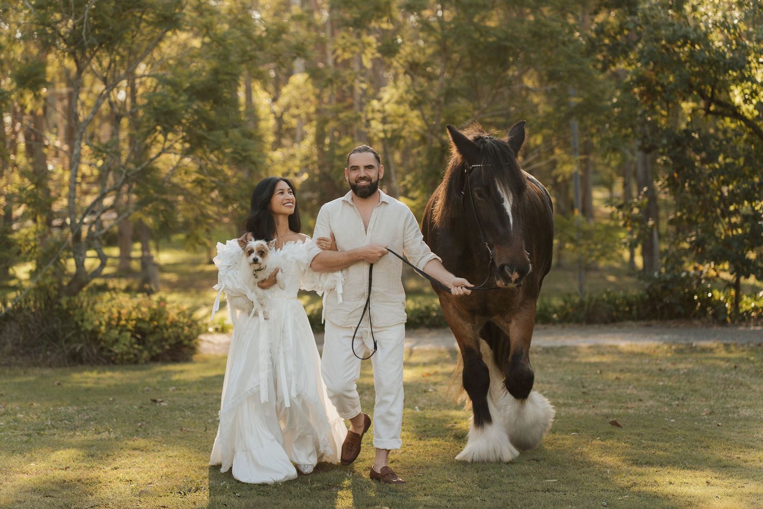 Couple in white walking with a brown horse and a mall dog in a green park setting in Sydney during an elopement photoshoot with a horse
