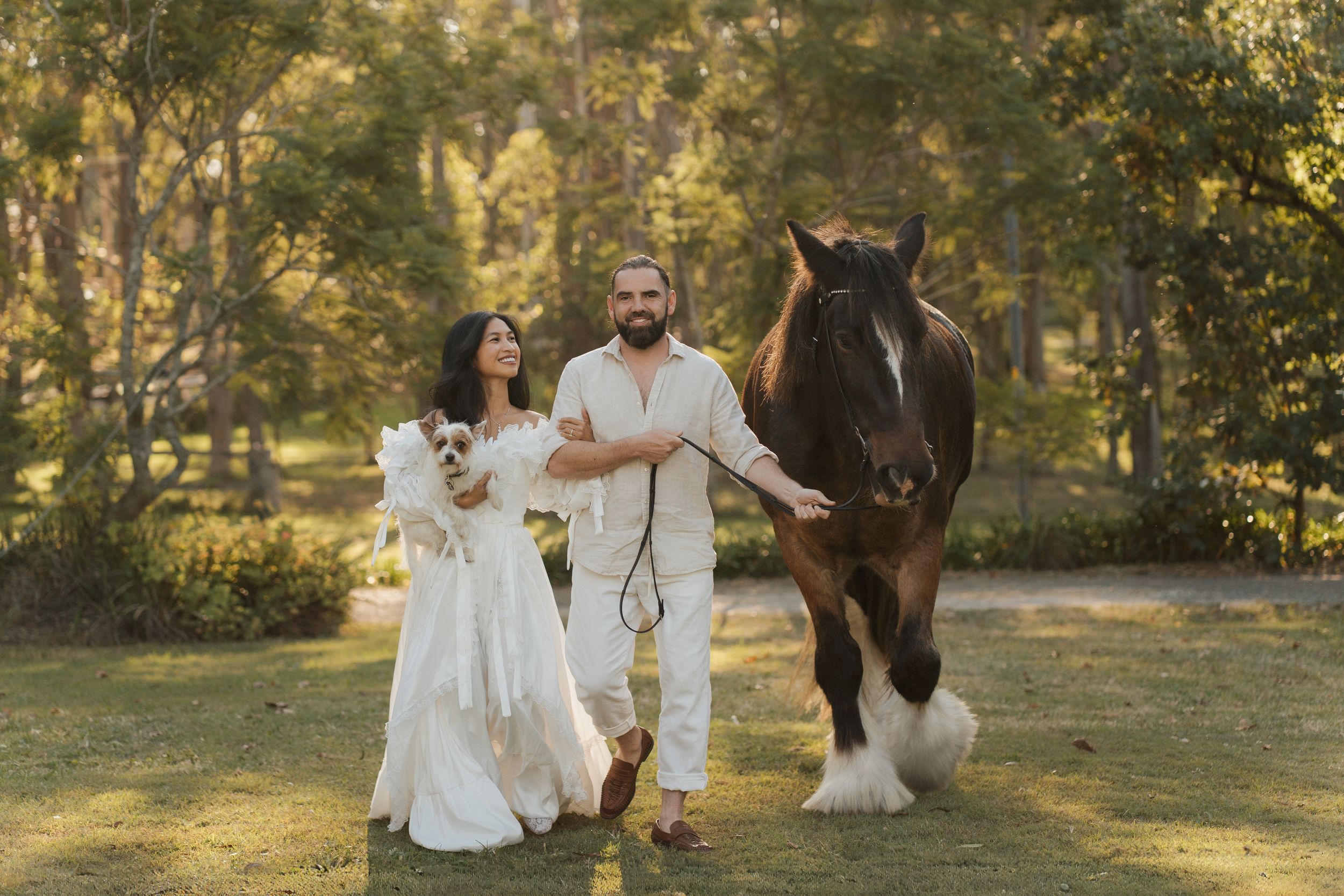 Couple in white walking with a brown horse and a mall dog in a green park setting in Sydney during an elopement photoshoot with a horse